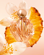 Cargar imagen en el visor de la galería, A faceted clear perfume bottle with a metallic gold cap, styled with a large pineapple slice and translucent white flower petals on a soft peach background.